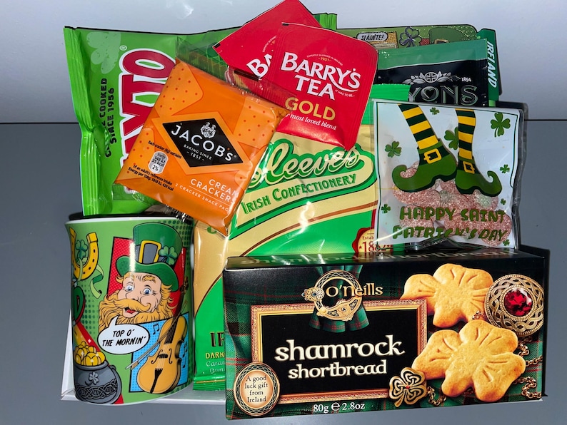 May include: A St. Patrick's Day gift basket with a green mug featuring a cartoon leprechaun, a bag of Jacobs Cream Crackers, a bag of Barry's Gold Tea, a bag of Reeves Irish Confectionery, a bag of O'Neill's Shamrock Shortbread, and a bag of green and gold candy.