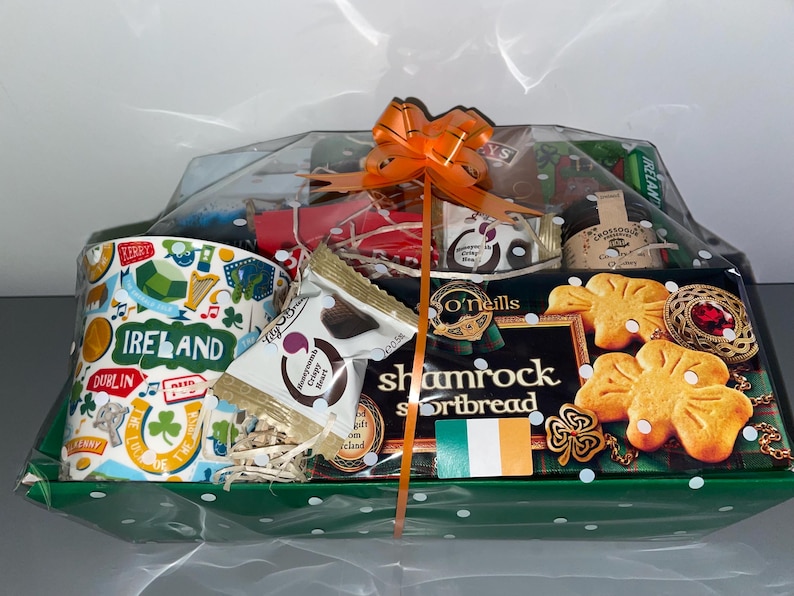 May include: A green and white gift basket filled with Irish themed treats. The basket includes a mug with the word "Ireland" on it, a box of chocolate covered crisps, a box of shortbread, and a jar of jam.