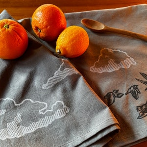 May include: Two gray tea towels with a white cloud design and a black bird design. The towels are folded and placed on a wooden table with three oranges and a wooden spoon.