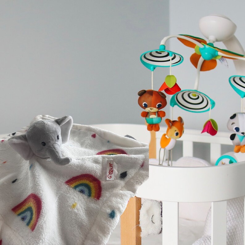 Puede incluir: Una manta blanca para beb&eacute; con una cabeza de elefante gris y detalles de arco&iacute;ris. Un m&oacute;vil para beb&eacute; con animales de peluche colgantes y formas coloridas es visible en el fondo. La manta est&aacute; sobre una cuna blanca.