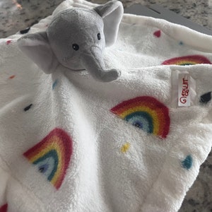 Puede incluir: Una manta blanca para beb&eacute; con una cabeza de elefante gris de peluche adjunta. La manta presenta arco&iacute;ris bordados y detalles coloridos. Una etiqueta con texto est&aacute; cosida en la manta.