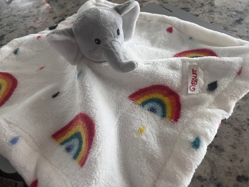 Puede incluir: Una manta blanca para beb&eacute; con una cabeza de elefante gris de peluche adjunta. La manta presenta arco&iacute;ris bordados y detalles coloridos. Una etiqueta con texto est&aacute; cosida en la manta.