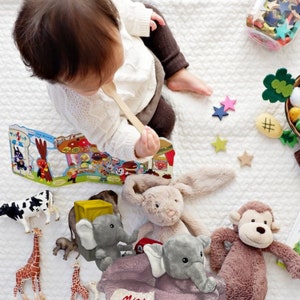 Puede incluir: Un beb&eacute; con una cuchara de madera est&aacute; rodeado de juguetes sobre una manta blanca. Los juguetes incluyen un conejito de peluche, un mono y un elefante, junto con figuras de animales y un libro. Tambi&eacute;n hay un tarro de confeti en forma de estrella.