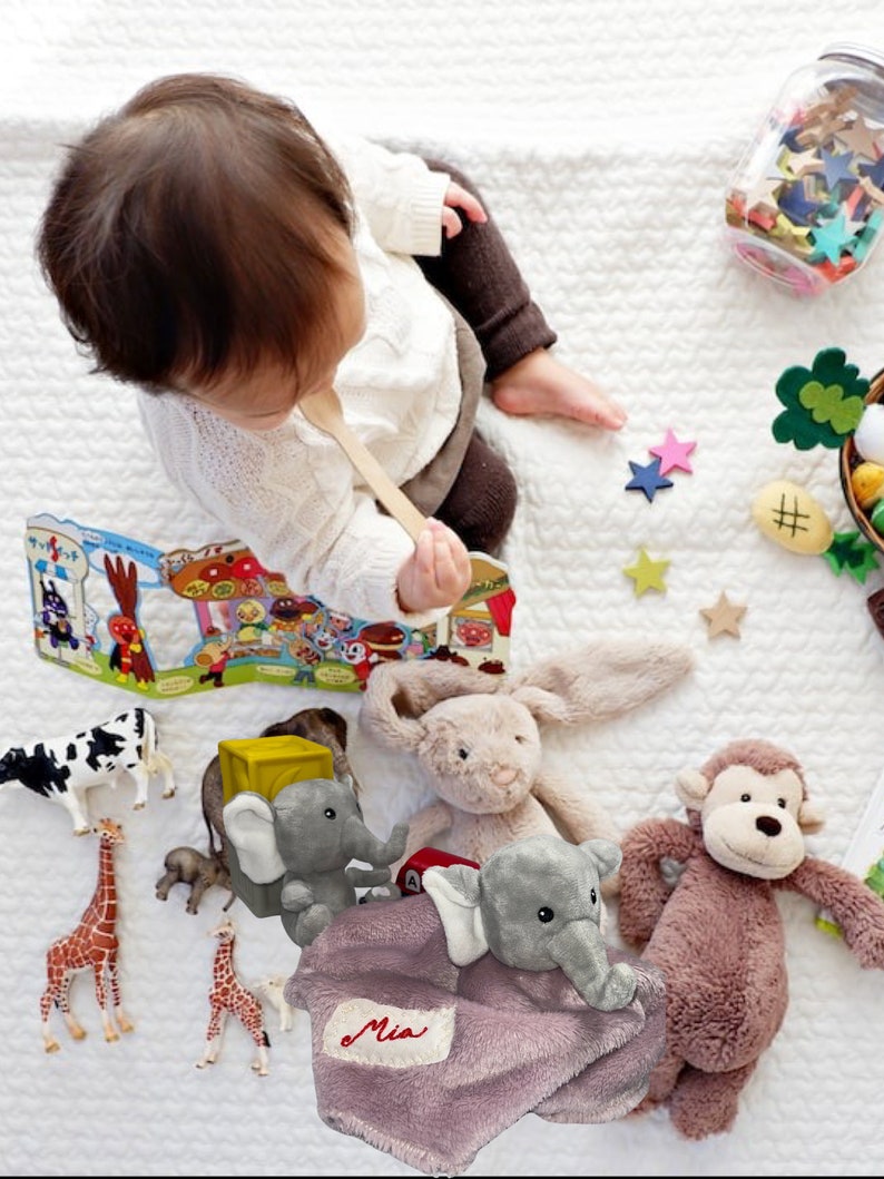 Puede incluir: Un beb&eacute; con una cuchara de madera est&aacute; rodeado de juguetes sobre una manta blanca. Los juguetes incluyen un conejito de peluche, un mono y un elefante, junto con figuras de animales y un libro. Tambi&eacute;n hay un tarro de confeti en forma de estrella.