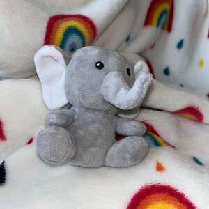 Puede incluir: Un peque&ntilde;o elefante de peluche gris con orejas y trompa blancas est&aacute; sentado sobre una manta blanca. La manta presenta un patr&oacute;n de arco&iacute;ris y gotas de lluvia en varios colores. El elefante tiene ojos negros y est&aacute; sentado.