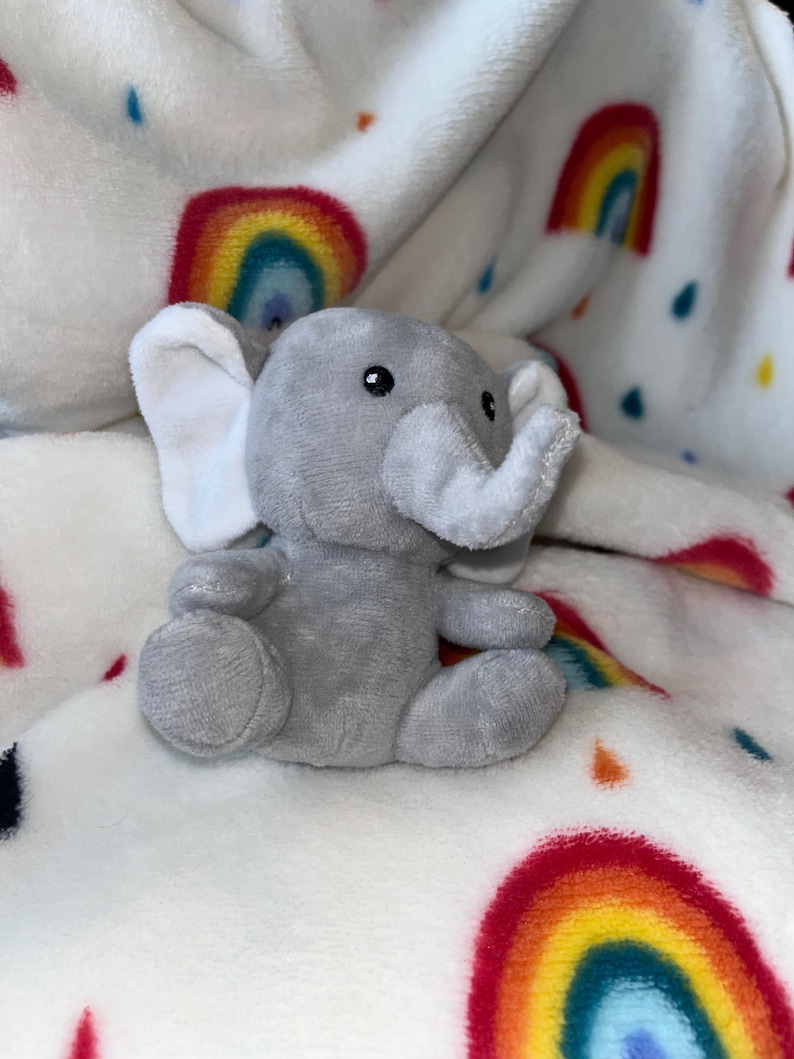 Puede incluir: Un peque&ntilde;o elefante de peluche gris con orejas y trompa blancas est&aacute; sentado sobre una manta blanca. La manta presenta un patr&oacute;n de arco&iacute;ris y gotas de lluvia en varios colores. El elefante tiene ojos negros y est&aacute; sentado.