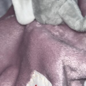 Puede incluir: Una manta de beb&eacute; de felpa con una cabeza de elefante gris y una manta suave de color lavanda. El elefante tiene ojos negros y orejas internas blancas. Una peque&ntilde;a etiqueta blanca con texto cursivo rojo est&aacute; cosida en la manta.