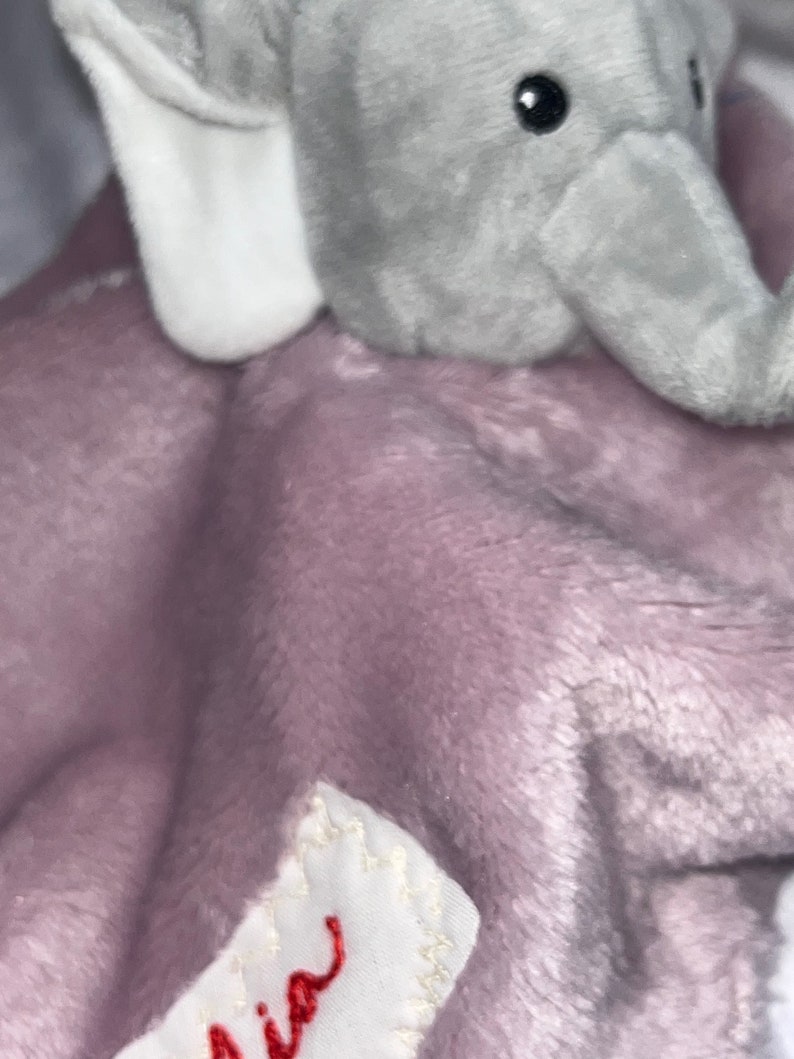 Puede incluir: Una manta de beb&eacute; de felpa con una cabeza de elefante gris y una manta suave de color lavanda. El elefante tiene ojos negros y orejas internas blancas. Una peque&ntilde;a etiqueta blanca con texto cursivo rojo est&aacute; cosida en la manta.