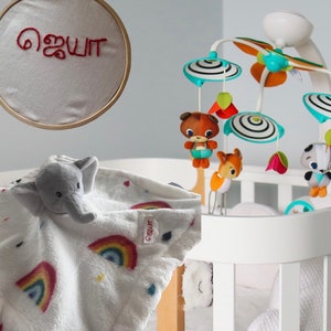 Puede incluir: Una cuna blanca con un m&oacute;vil con figuras de animales y elementos decorativos. Una manta blanca con un dise&ntilde;o de arco&iacute;ris y una cabeza de elefante est&aacute; sobre la cuna. Tambi&eacute;n se ve un aro bordado con el nombre "ஜெயா" en rojo.