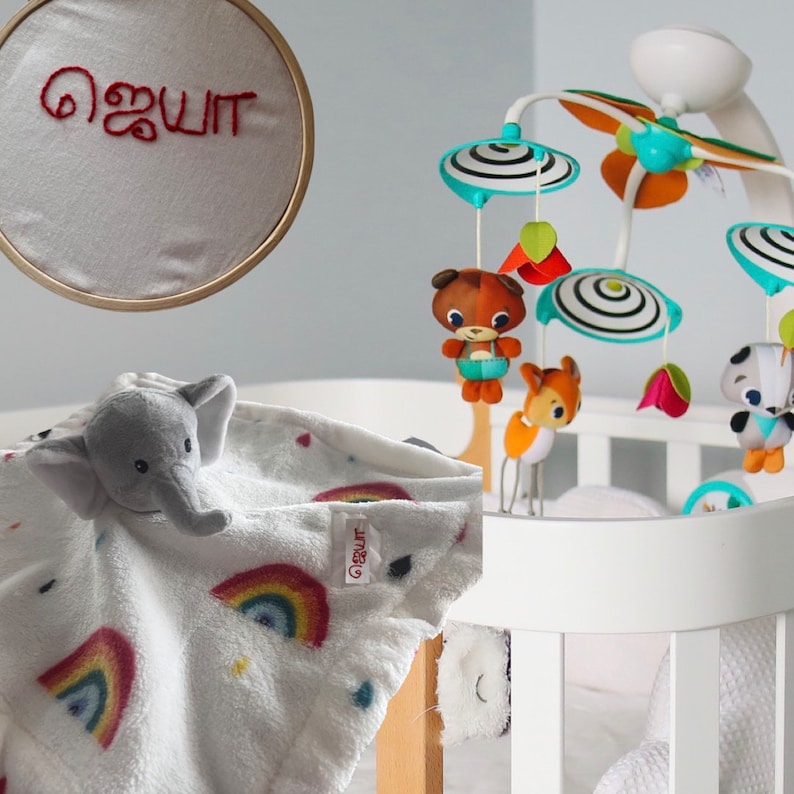 Puede incluir: Una cuna blanca con un m&oacute;vil con figuras de animales y elementos decorativos. Una manta blanca con un dise&ntilde;o de arco&iacute;ris y una cabeza de elefante est&aacute; sobre la cuna. Tambi&eacute;n se ve un aro bordado con el nombre "ஜெயா" en rojo.