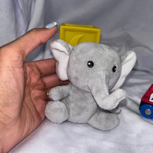 Puede incluir: Un juguete de elefante de peluche gris con orejas internas blancas y ojos negros. Un bloque amarillo con un dise&ntilde;o curvo est&aacute; detr&aacute;s del elefante. Un tren de juguete rojo y azul es visible en el fondo.