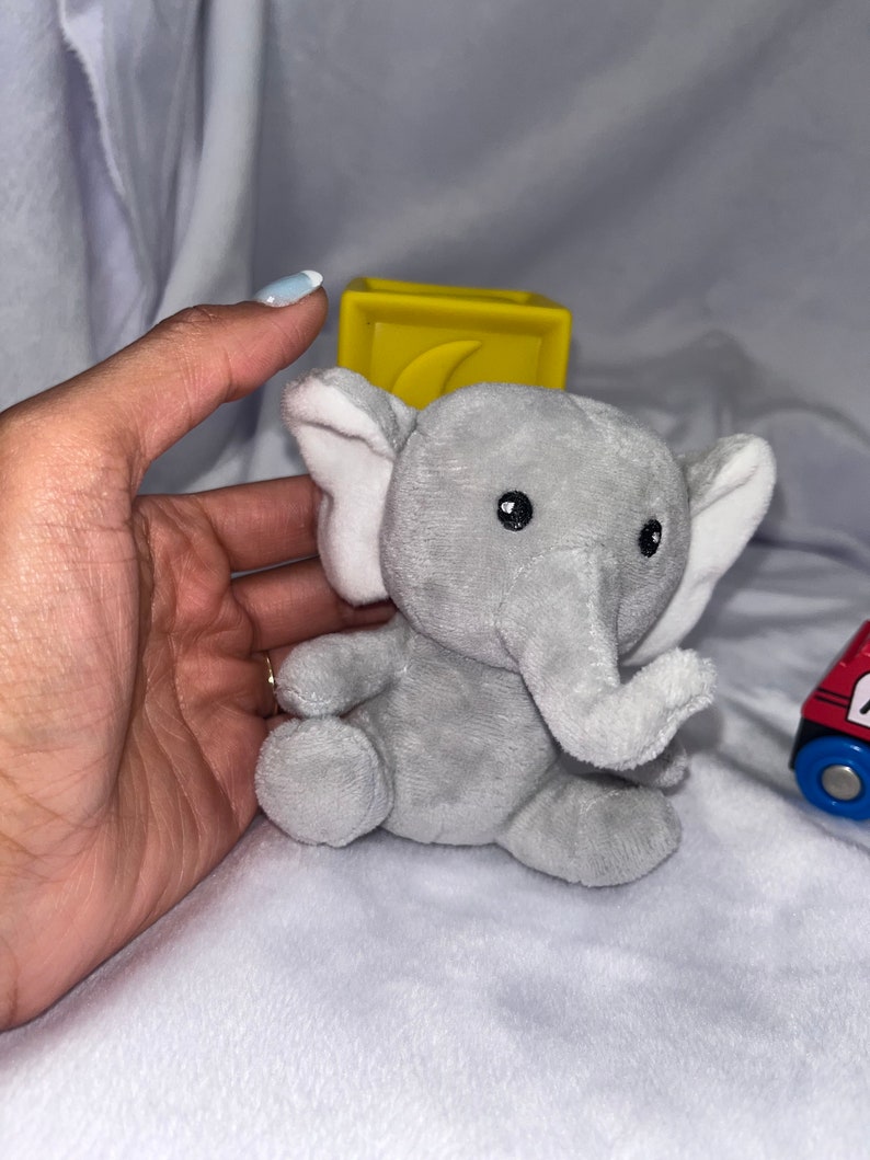 Puede incluir: Un juguete de elefante de peluche gris con orejas internas blancas y ojos negros. Un bloque amarillo con un dise&ntilde;o curvo est&aacute; detr&aacute;s del elefante. Un tren de juguete rojo y azul es visible en el fondo.
