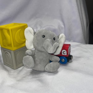 Puede incluir: Un juguete de elefante de peluche gris con orejas blancas est&aacute; sentado junto a un vag&oacute;n de tren de juguete rojo y azul. Un bloque de juguete amarillo y gris con un dise&ntilde;o de luna y arco tambi&eacute;n est&aacute; en la imagen. Los juguetes est&aacute;n sobre una superficie blanca texturizada.