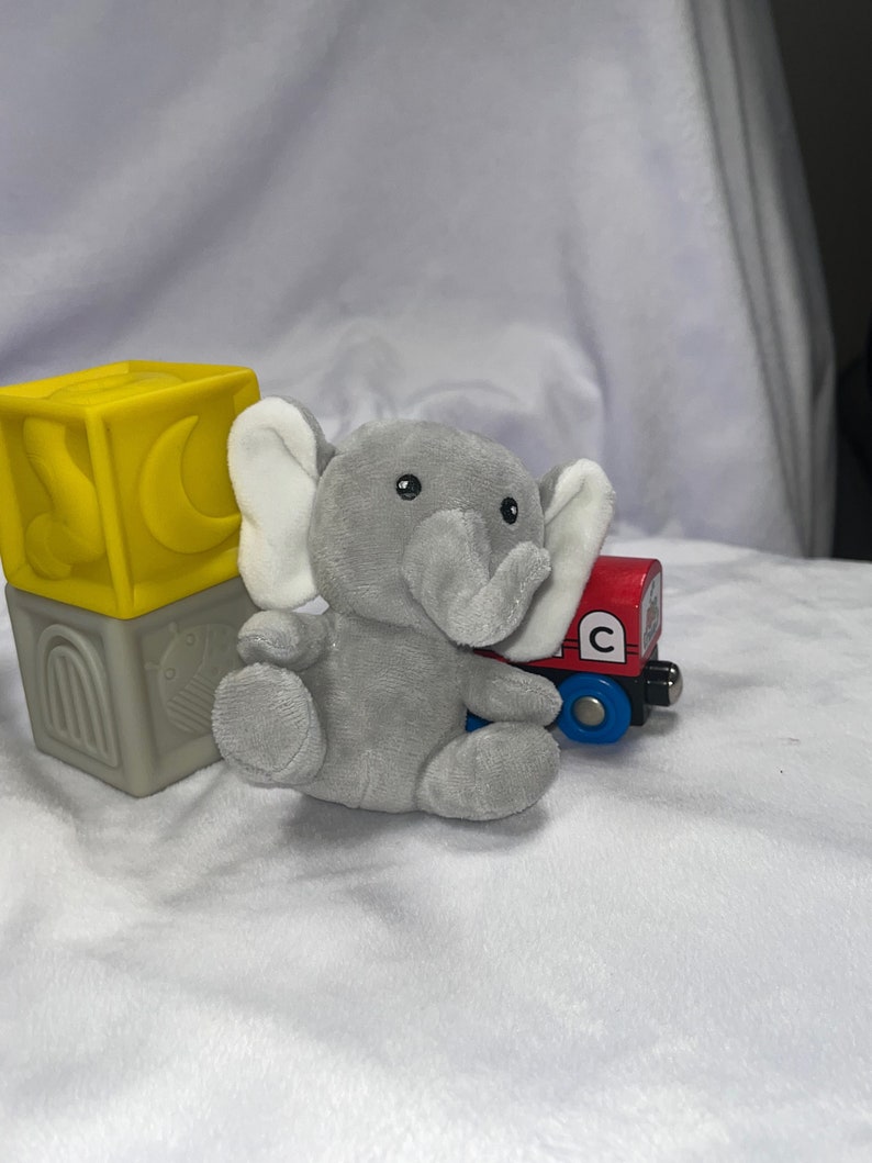 Puede incluir: Un juguete de elefante de peluche gris con orejas blancas est&aacute; sentado junto a un vag&oacute;n de tren de juguete rojo y azul. Un bloque de juguete amarillo y gris con un dise&ntilde;o de luna y arco tambi&eacute;n est&aacute; en la imagen. Los juguetes est&aacute;n sobre una superficie blanca texturizada.