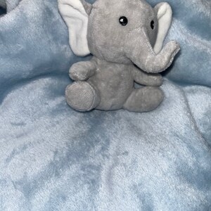 Puede incluir: Un peque&ntilde;o elefante de peluche gris con orejas internas blancas est&aacute; sentado sobre una manta de felpa azul claro. El elefante tiene ojos negros y una trompa. La manta tiene una textura suave y difusa.