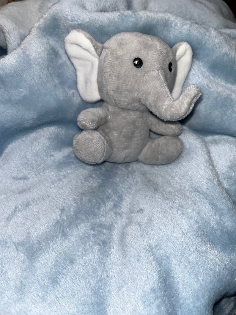 Puede incluir: Un peque&ntilde;o elefante de peluche gris con orejas internas blancas est&aacute; sentado sobre una manta de felpa azul claro. El elefante tiene ojos negros y una trompa. La manta tiene una textura suave y difusa.