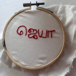 Puede incluir: Un bastidor de bordado con la palabra roja "ஜெயா" bordada sobre tela blanca. El bastidor es de madera y tiene un mecanismo de apriete met&aacute;lico. Un trozo de tela blanca est&aacute; detr&aacute;s del bastidor.