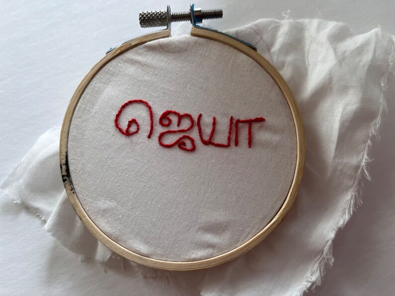 Puede incluir: Un bastidor de bordado con la palabra roja "ஜெயா" bordada sobre tela blanca. El bastidor es de madera y tiene un mecanismo de apriete met&aacute;lico. Un trozo de tela blanca est&aacute; detr&aacute;s del bastidor.