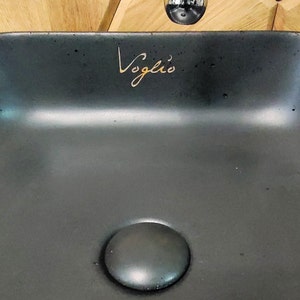 Könnte beinhalten: Ein schwarzes Keramikwaschbecken mit einem runden Abfluss und dem Wort "Voglìo" in goldenen Buchstaben.