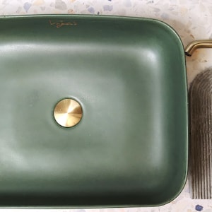 Könnte beinhalten: Ein rechteckiges, olivgrünes Waschbecken mit einem goldfarbenen Abfluss und einer passenden Armatur. Ein graues, bogenförmiges Dekorationselement und eine goldfarbene Armatur befinden sich rechts vom Waschbecken. Das Waschbecken hat den Schriftzug "byjess" in Gold.