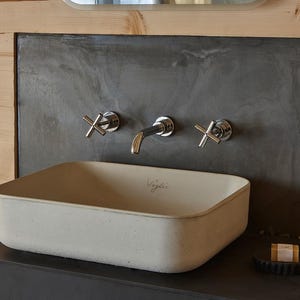 May include: A modern bathroom sink with chrome fixtures. The rectangular, light-colored sink is mounted on a dark countertop against a dark gray wall. The wall-mounted faucet and cross-shaped handles add a touch of elegance. The word "Vigle" is written on the sink.