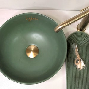 Könnte beinhalten: Ein grünes Keramikwaschbecken mit einem goldenen Abfluss und dem Wort "Vogue" in goldenen Lettern. Das Waschbecken wird von einem passenden grünen Tablett mit einem dekorativen Muster begleitet.