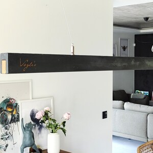 May include: A rectangular, dark grey pendant light fitting with the word "Voglio" in a copper-coloured script. The light has a gold-coloured end cap and is suspended from the ceiling by a thin white cord. The fitting is in a modern living space.