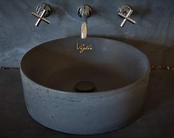 May include: A dark gray, round concrete bathroom sink with a matte finish. The sink has a black drain and the word "Voglio" in gold script. Chrome faucets with cross handles are mounted above the sink.