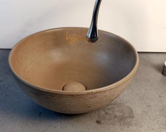 May include: A round, beige concrete vessel sink with a smooth interior and a textured exterior surface. The sink features a central drain plug and a subtle, cursive gold logo near the rim. A polished chrome faucet stands to the right side.