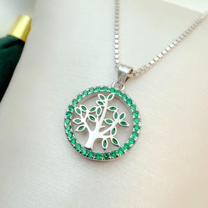 May include: Silver-toned necklace featuring a circular pendant with a tree design. The tree is surrounded by a ring of small, green gemstones. The necklace has a delicate chain and a simple clasp.