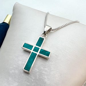 Puede incluir: Un colgante de cruz plateada con incrustaciones de color turquesa, suspendido de una fina cadena de plata. El diseño de la cruz presenta una forma clásica con detalles rectangulares turquesa. El collar se exhibe sobre una superficie blanca.
