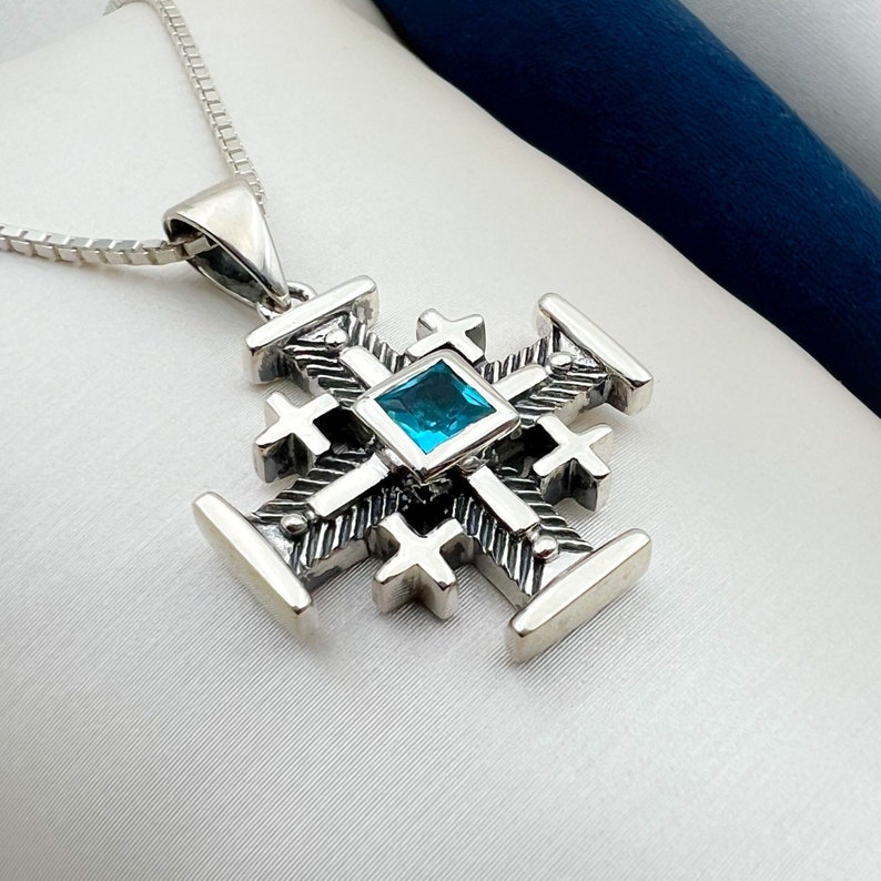 Pu&ograve; includere: Una collana con un ciondolo in argento a forma di croce dettagliata con una pietra preziosa blu al centro.