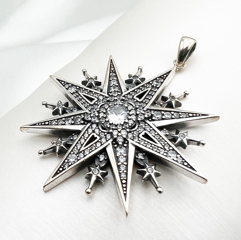 May include: A silver pendant in the shape of a large, eight-pointed star with a smaller star at each point. The star is covered in small, clear crystals.
