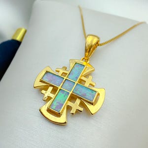 Puede incluir: Un collar con colgante dorado con un diseño de cruz de Jerusalén. La cruz tiene un cuadrado central con incrustaciones iridiscentes de ópalo azul y verde. La cadena y el colgante son de metal dorado.