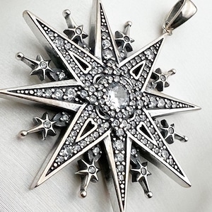 May include: A silver pendant in the shape of a star with a large clear crystal in the center. The star is surrounded by smaller crystals and has a detailed design.