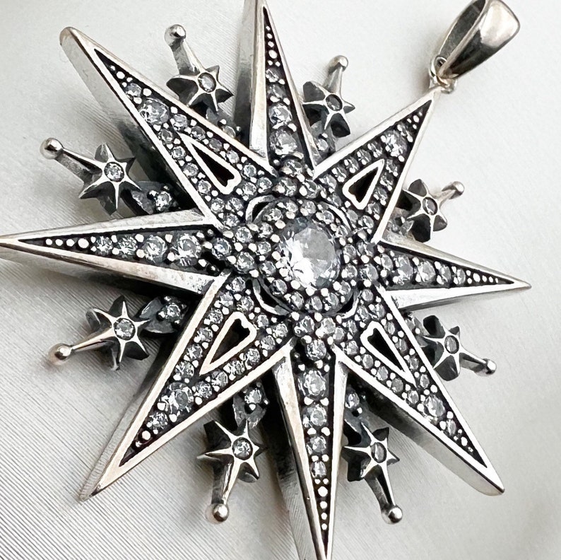 May include: A silver pendant in the shape of a star with a large clear crystal in the center. The star is surrounded by smaller crystals and has a detailed design.