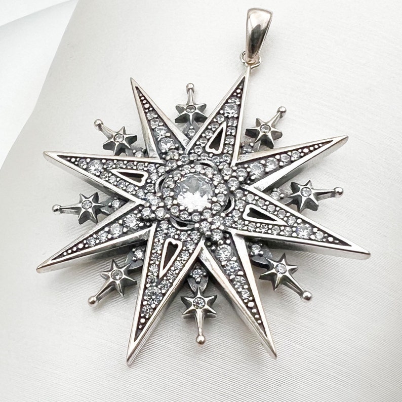 May include: A silver pendant in the shape of a star with a large clear crystal in the center. The star has eight points and is surrounded by smaller crystals.
