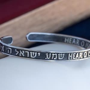Può includere: Un bracciale a polsino color argento con testo ebraico e inglese inciso. Il bracciale aperto è esposto su una superficie bianca. Il testo include la frase "Hear O Israel".