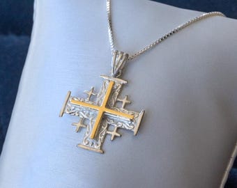 May include: A silver-toned Jerusalem cross pendant with a gold-colored center cross, suspended from a delicate chain. The cross features intricate detailing and is displayed on a light gray surface.