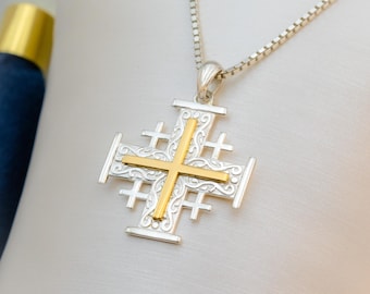 May include: A silver pendant necklace featuring a Jerusalem cross design with a gold-colored cross in the center. The cross has decorative filigree patterns and is suspended from a delicate silver chain. The pendant is displayed against a white background.