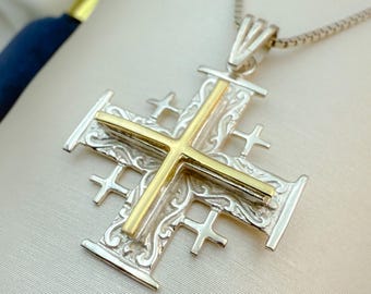 May include: A silver Jerusalem cross pendant with a gold-colored cross in the center. The pendant has an ornate design and is suspended from a silver chain. The cross is a religious symbol.