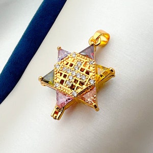 May include: Gold-toned star pendant with a central geometric design encrusted with small, clear crystals. The star points feature colorful, triangular gemstones in shades of pink, yellow, and green. The pendant is displayed on a white surface.