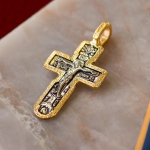 Puede incluir: Un colgante de cruz de metal dorado con un relieve central plateado de un crucifijo. La cruz tiene un borde texturizado y ornamentado, e incluye texto en cir&iacute;lico grabado e iconograf&iacute;a religiosa en el centro plateado.