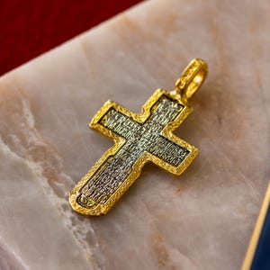 Puede incluir: Un colgante de cruz en tono dorado con un panel central de metal oscuro texturizado, grabado con intrincada escritura cir&iacute;lica. La cruz tiene un borde dorado pulido en relieve con un patr&oacute;n decorativo y una peque&ntilde;a anilla superior.