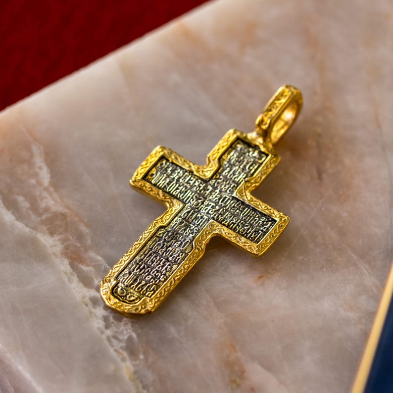 Puede incluir: Un colgante de cruz en tono dorado con un panel central de metal oscuro texturizado, grabado con intrincada escritura cir&iacute;lica. La cruz tiene un borde dorado pulido en relieve con un patr&oacute;n decorativo y una peque&ntilde;a anilla superior.