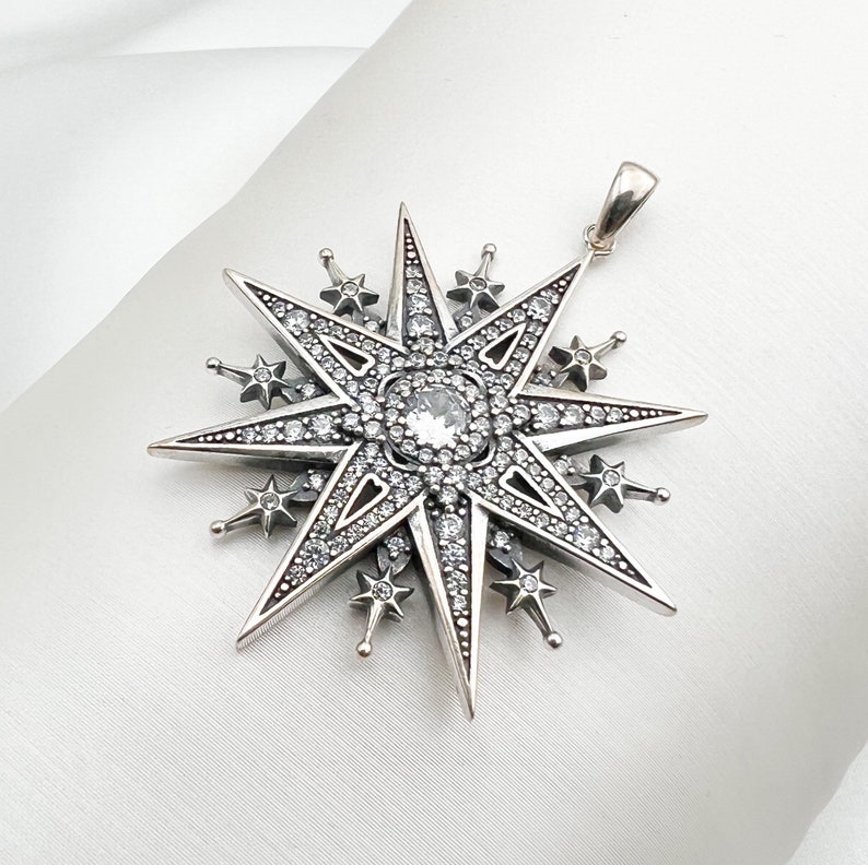 May include: A silver pendant in the shape of a large, eight-pointed star with smaller stars around the perimeter. The star is encrusted with clear rhinestones.