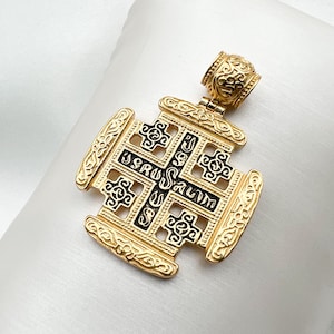 Puede incluir: Un colgante dorado con un diseño de cruz de Jerusalén en esmalte negro. La cruz es intrincada y presenta la inscripción "Jerusalén" en esmalte negro.