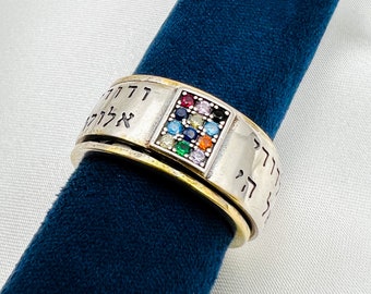 May include: A silver ring with a band of Hebrew text and a square inlay of colorful gemstones. The ring is on a blue velvet surface.