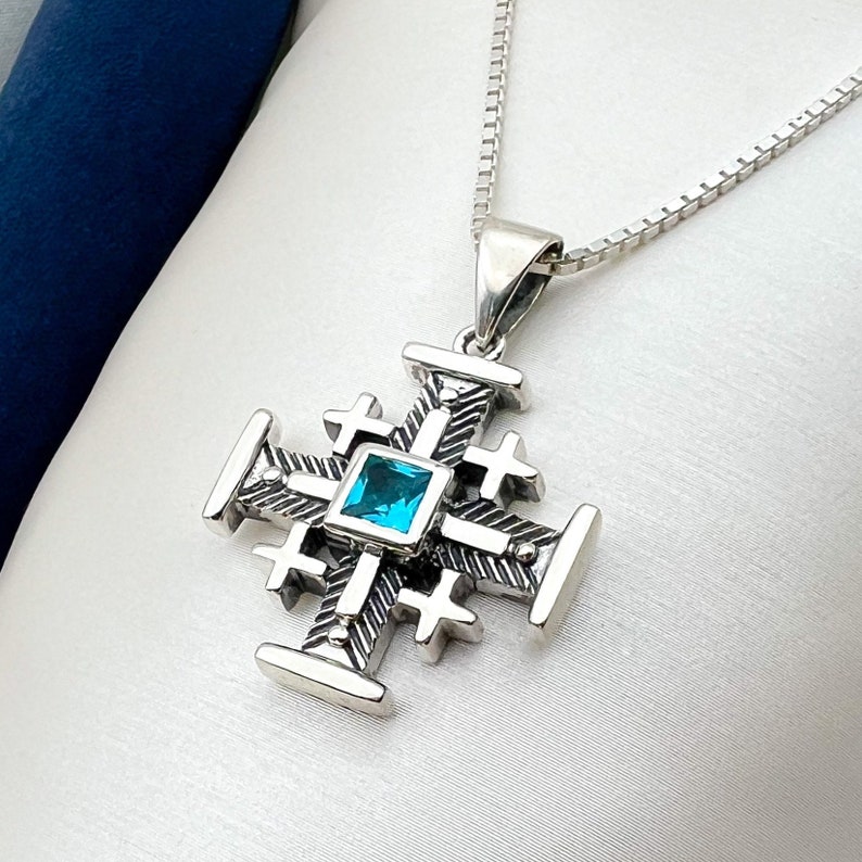Pu&ograve; includere: Una collana con ciondolo in argento a forma di croce dettagliata con una pietra preziosa blu al centro.
