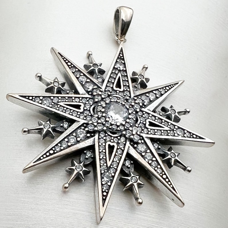 May include: A silver pendant in the shape of a eight-pointed star with a large clear crystal in the center. The star is decorated with smaller crystals around the edges.